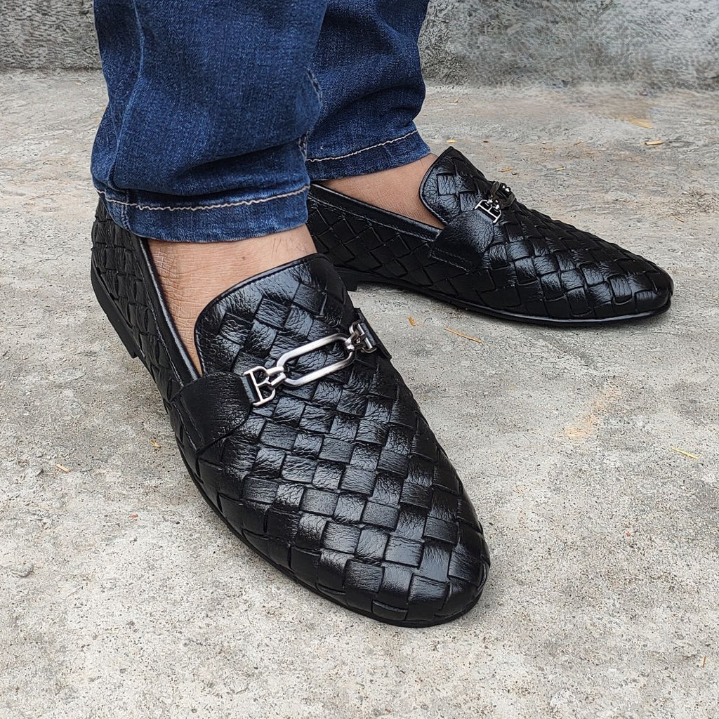 Black woven loafers with a silver buckle worn by a person on a concrete surface.