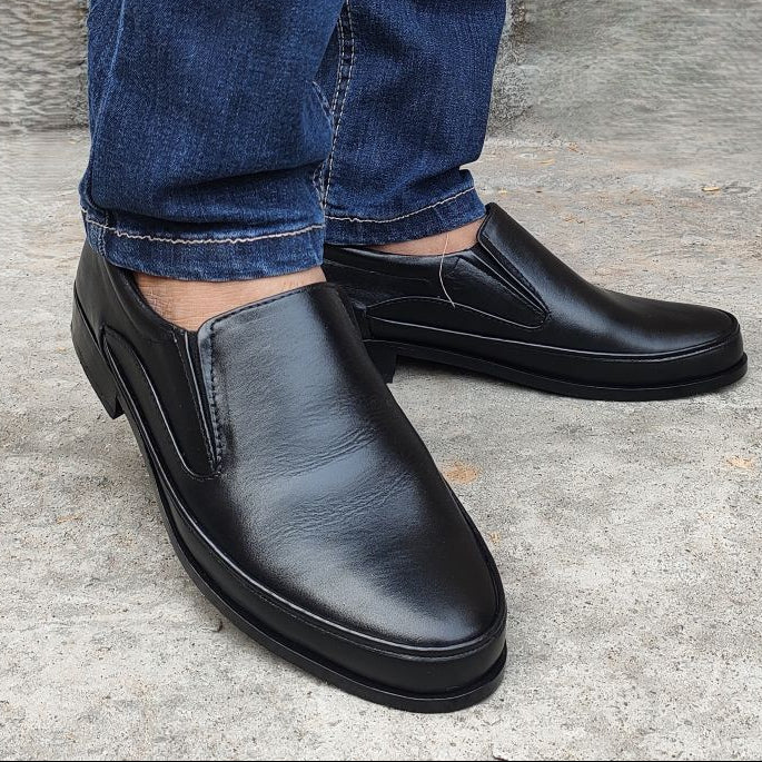 Black slip-on shoes worn with blue jeans on a concrete floor.