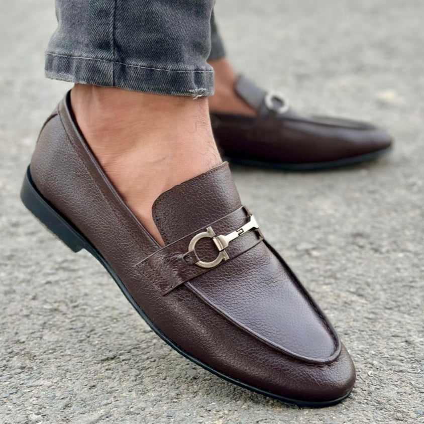 Brown loafers with a decorative buckle worn on a gray pavement.
