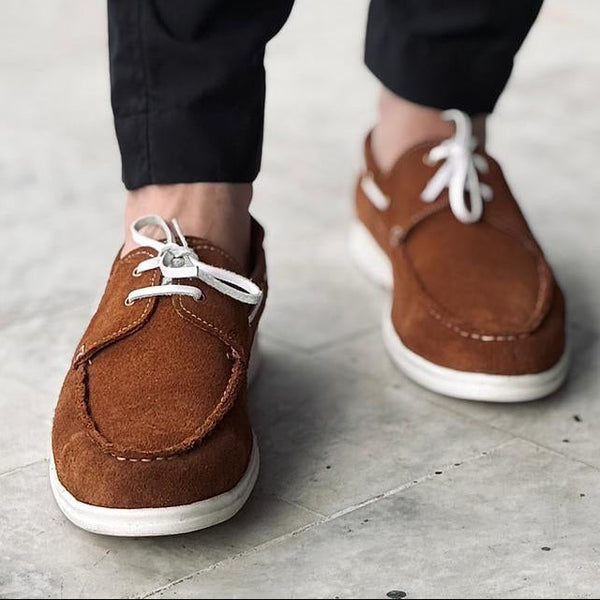 Brown boat shoes with white laces worn with black pants on a light gray pavement.